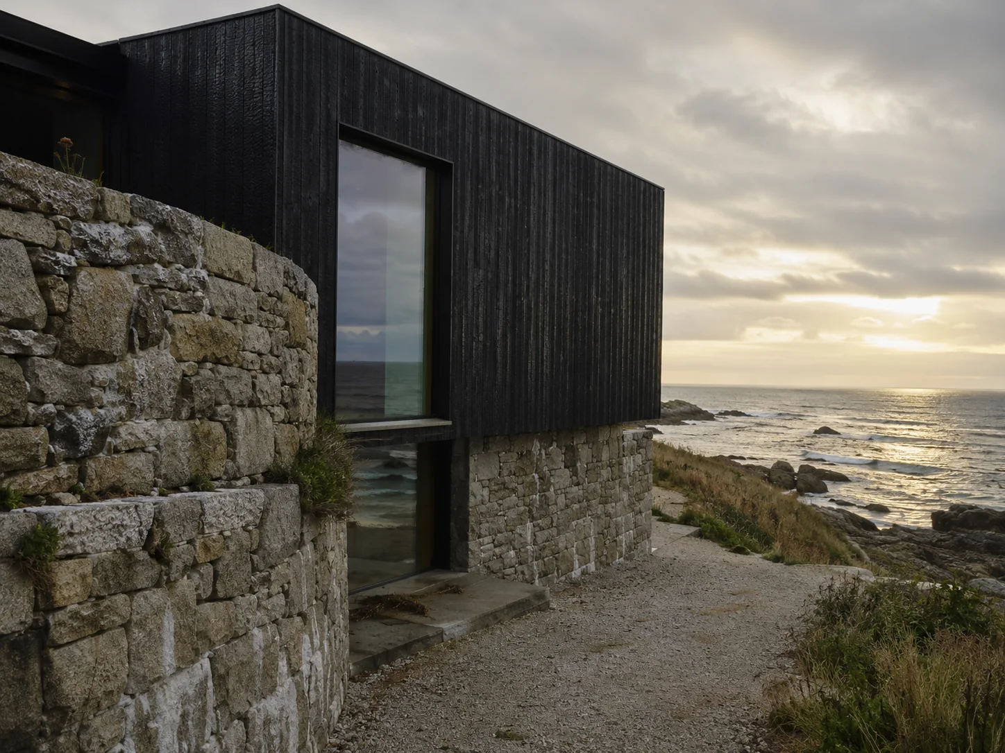 Maison côtière en bois brûlé noir posée sur un socle de pierre, face à la mer.
